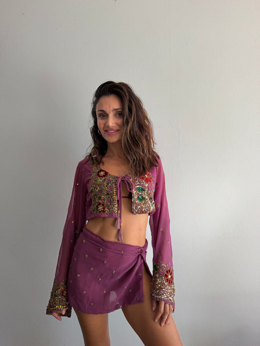 a two-piece bohemian-style outfit consisting of a sheer long-sleeve crop top and a matching mini skirt in a dusty purple shade, both heavily decorated with colorful sequins and embroidery.